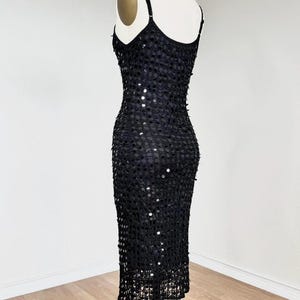May include: A black sequined dress on a mannequin. The dress features thin spaghetti straps and a fitted silhouette. The dress is covered in small, round sequins, creating a shimmering effect. The hemline falls to the mid-calf.