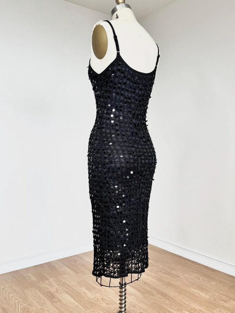 May include: A black sequined dress on a mannequin. The dress features thin spaghetti straps and a fitted silhouette. The dress is covered in small, round sequins, creating a shimmering effect. The hemline falls to the mid-calf.