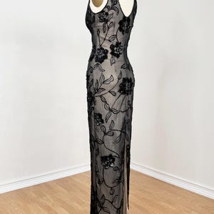 May include: A black evening gown with floral embellishments. The sleeveless dress features a sheer overlay adorned with black sequins in a floral pattern. The gown has a V-neckline and a high slit on the side, displayed on a mannequin.