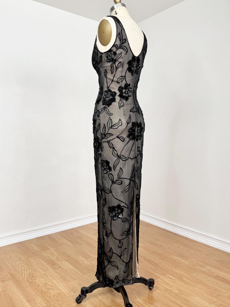 May include: A black evening gown with floral embellishments. The sleeveless dress features a sheer overlay adorned with black sequins in a floral pattern. The gown has a V-neckline and a high slit on the side, displayed on a mannequin.