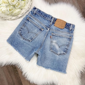 May include: Light blue denim shorts with a frayed hem. The shorts feature classic five-pocket styling, a button and zipper closure, and the Levi's brand patch on the back. The shorts are displayed on a white fluffy surface.