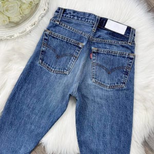 May include: Blue denim jeans with classic five-pocket styling. The jeans feature a faded wash, a red tag on the back pocket, and a white label on the waistband. The jeans are displayed on a white, fluffy surface.