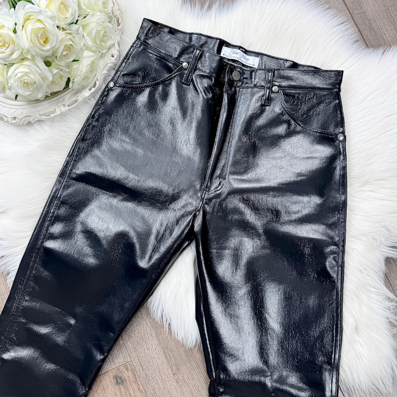 May include: Black, high-waisted trousers with a shiny, leather-look finish. The trousers feature a classic five-pocket design with silver rivets and a button closure. The trousers are displayed on a white, fluffy surface.