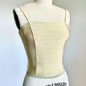 May include: A gold-colored bustier top with thin straps and a square neckline. The bodice has a textured appearance and is fitted, with a decorative trim along the bottom edge. The top is displayed on a mannequin.