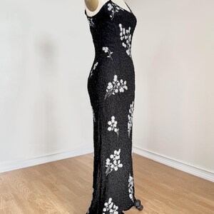 May include: A black evening gown with spaghetti straps, adorned with white floral embroidery. The dress is form-fitting and floor-length, displayed on a mannequin. The fabric appears to be embellished with small beads.