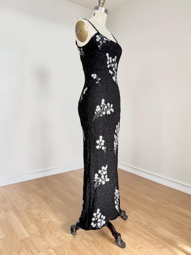 May include: A black evening gown with spaghetti straps, adorned with white floral embroidery. The dress is form-fitting and floor-length, displayed on a mannequin. The fabric appears to be embellished with small beads.
