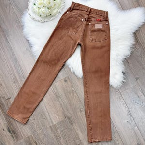May include: Brown denim jeans with a classic five-pocket design. The jeans feature a straight leg cut and a small leather patch on the back pocket. The Wrangler brand label is visible.