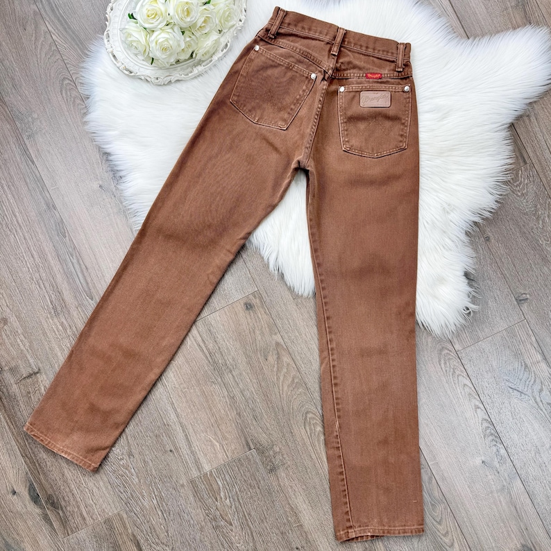 May include: Brown denim jeans with a classic five-pocket design. The jeans feature a straight leg cut and a small leather patch on the back pocket. The Wrangler brand label is visible.
