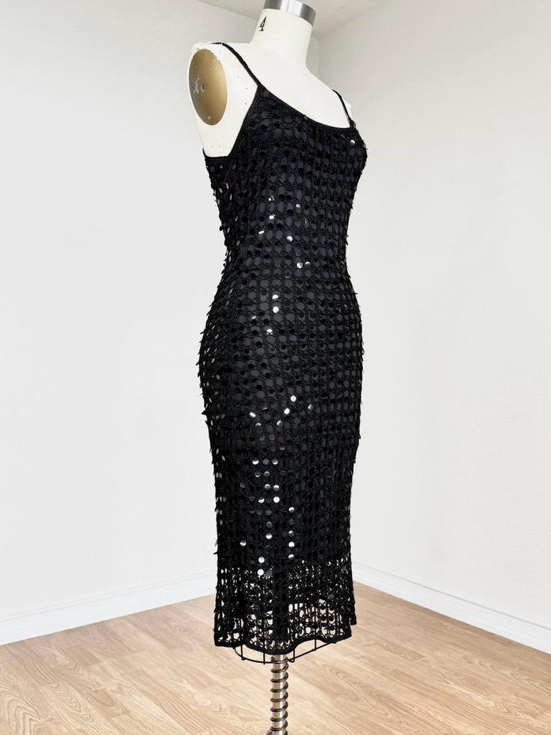 May include: A black sequined dress with spaghetti straps, displayed on a mannequin. The dress is knee-length and features a fitted silhouette. The sequins are evenly spaced, creating a shimmering effect. The dress is suitable for formal events.