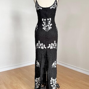 May include: A black beaded evening gown with white floral embroidery. The dress features a fitted silhouette, spaghetti straps, and a high slit in the front. The gown is displayed on a dress form.