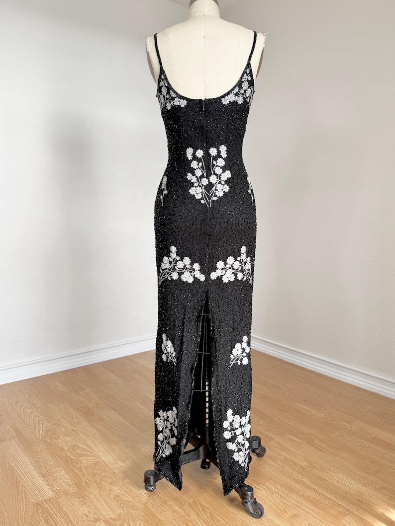 May include: A black beaded evening gown with white floral embroidery. The dress features a fitted silhouette, spaghetti straps, and a high slit in the front. The gown is displayed on a dress form.