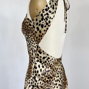 May include: A sleeveless dress featuring a tan and black leopard print pattern. The garment has a high neckline, a deep open back design, and thin straps that tie at the nape of the neck, displayed on a mannequin to show the side profile.