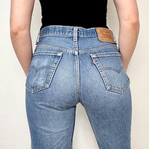 May include: Medium-wash blue denim jeans featuring a high-rise waist, two back pockets with signature arcuate stitching, a small red tab on the right pocket, and a tan leather patch on the waistband with the Levi Strauss & Co. brand logo.