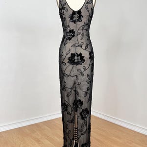 May include: A black evening gown with a floral design. The sleeveless dress features a V-neckline and a floor-length skirt with a slit in the back. The dress is adorned with black sequins and beads, creating a shimmering effect. The dress is displayed on a mannequin.