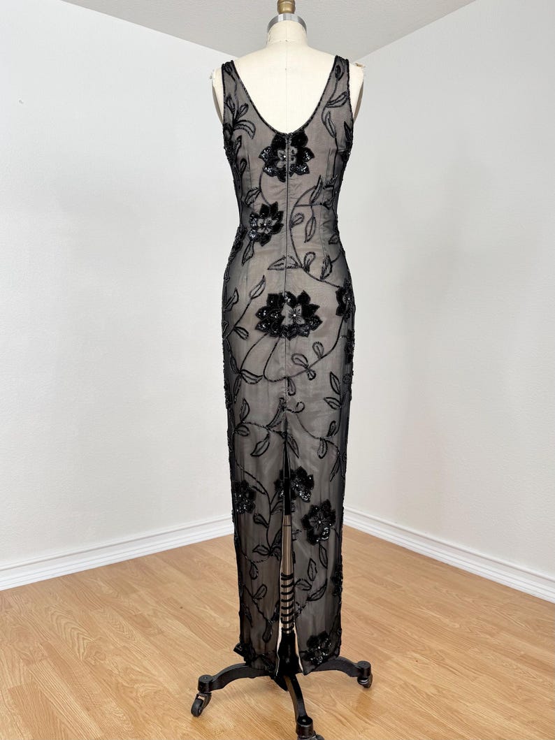 May include: A black evening gown with a floral design. The sleeveless dress features a V-neckline and a floor-length skirt with a slit in the back. The dress is adorned with black sequins and beads, creating a shimmering effect. The dress is displayed on a mannequin.