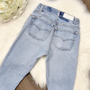 May include: Light blue denim jeans with classic five-pocket styling. The jeans feature a faded wash, a straight leg, and the iconic Levi's back pocket design. A white label is visible on the back waistband. The jeans are displayed on a white faux fur surface.
