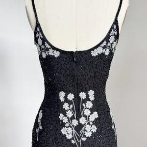 May include: Black beaded evening dress with spaghetti straps and a floral design. The dress features white floral embroidery on the bodice and back, with a fitted silhouette. The dress is displayed on a mannequin.
