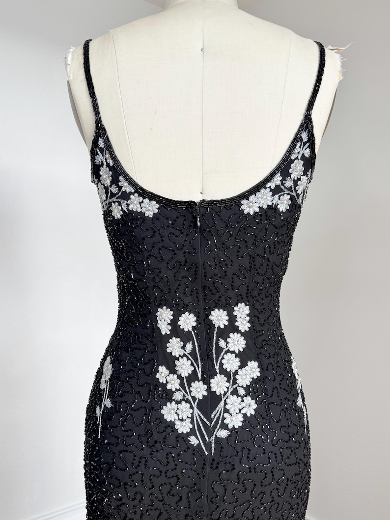 May include: Black beaded evening dress with spaghetti straps and a floral design. The dress features white floral embroidery on the bodice and back, with a fitted silhouette. The dress is displayed on a mannequin.