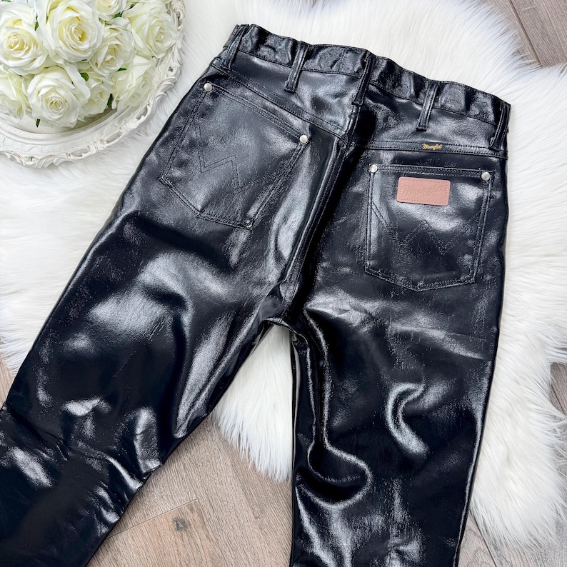 May include: Black, shiny Wrangler jeans with a high waist and classic five-pocket design. The back pockets feature the iconic Wrangler 'W' stitching. A brown leather patch is visible on the back. The trousers are displayed on a white fur-like surface.