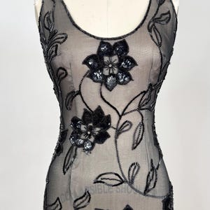 May include: A sleeveless black evening gown with floral embellishments. The dress features a sheer overlay adorned with beaded and sequined floral designs. The flowers are concentrated on the bodice and cascade down the dress, creating a glamorous look.