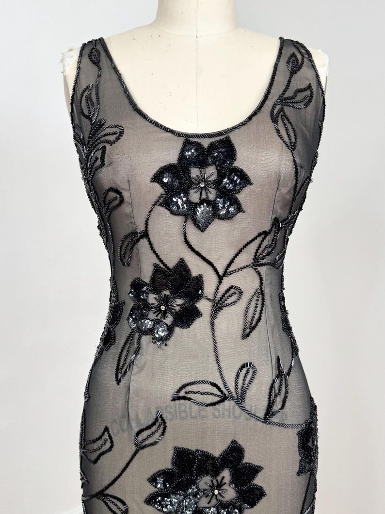 May include: A sleeveless black evening gown with floral embellishments. The dress features a sheer overlay adorned with beaded and sequined floral designs. The flowers are concentrated on the bodice and cascade down the dress, creating a glamorous look.