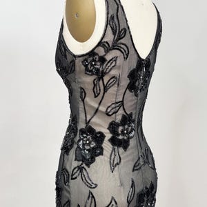 May include: A black evening gown with floral beadwork. The sleeveless dress features a sheer overlay adorned with intricate black beaded flowers and leaves. The gown is displayed on a mannequin, showcasing its elegant silhouette and design.