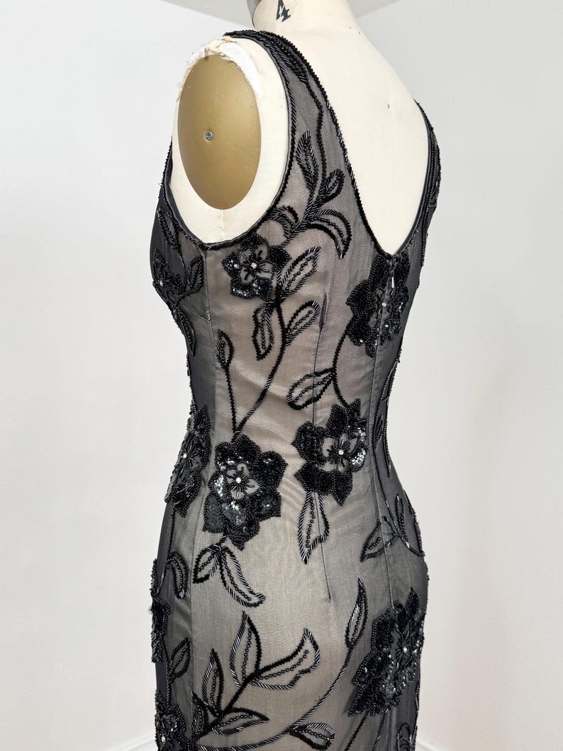 May include: A black evening gown with floral beadwork. The sleeveless dress features a sheer overlay adorned with intricate black beaded flowers and leaves. The gown is displayed on a mannequin, showcasing its elegant silhouette and design.