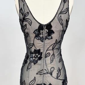 May include: A black evening dress with a V-neck and floral embellishments. The dress features a sheer overlay adorned with beaded floral patterns and sequin accents. The dress is displayed on a mannequin.