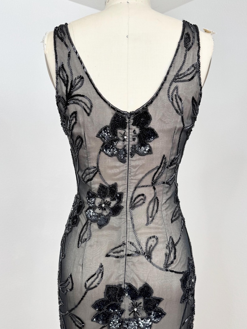 May include: A black evening dress with a V-neck and floral embellishments. The dress features a sheer overlay adorned with beaded floral patterns and sequin accents. The dress is displayed on a mannequin.