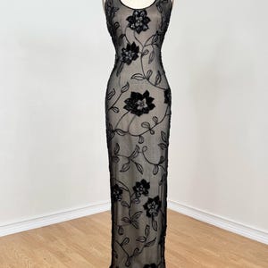 May include: A black, floor-length evening gown with a sheer overlay, adorned with floral embroidery. The sleeveless dress features a scoop neckline and is displayed on a mannequin. The floral design is concentrated on the bodice and cascades down the dress.