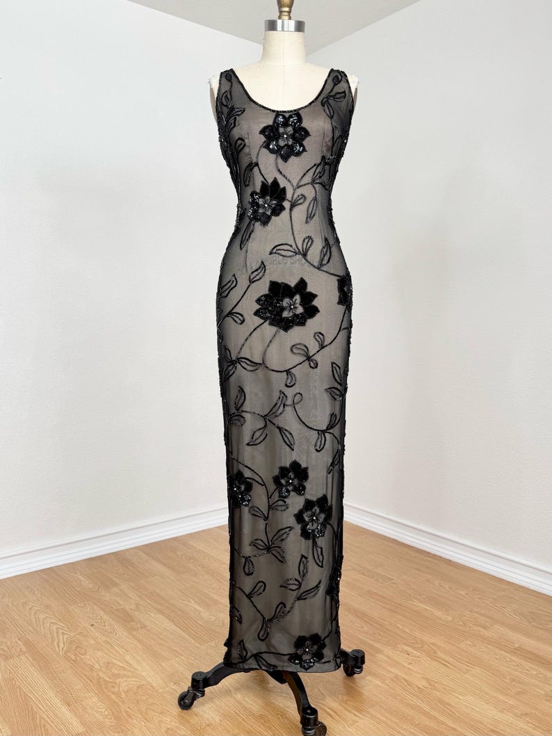 May include: A black, floor-length evening gown with a sheer overlay, adorned with floral embroidery. The sleeveless dress features a scoop neckline and is displayed on a mannequin. The floral design is concentrated on the bodice and cascades down the dress.