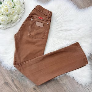 May include: Brown denim jeans with a classic five-pocket design. Features a leather patch and a red logo tag. The jeans are laid flat on a white fluffy surface.