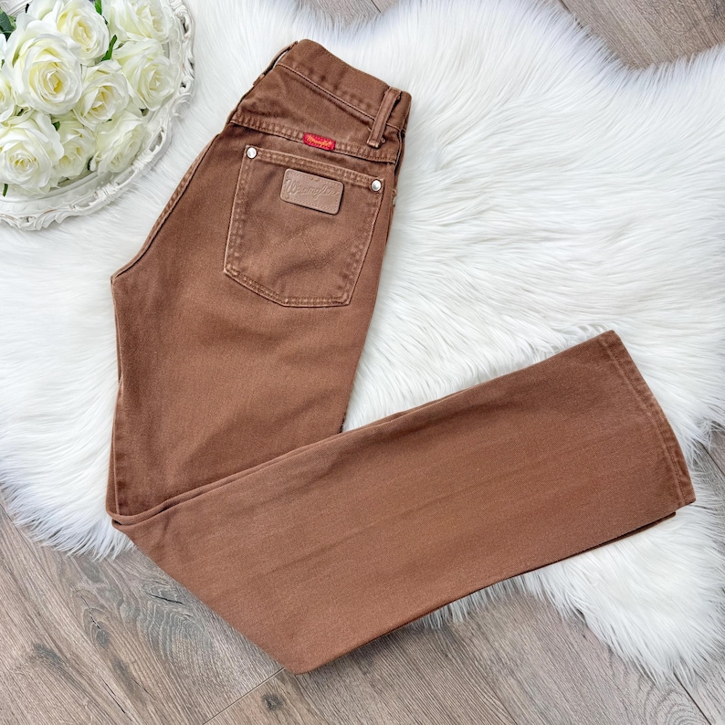May include: Brown denim jeans with a classic five-pocket design. Features a leather patch and a red logo tag. The jeans are laid flat on a white fluffy surface.