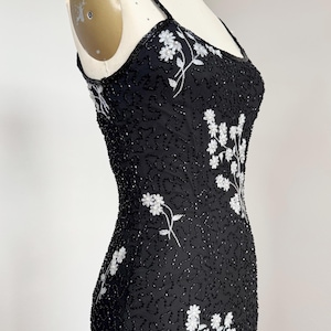 May include: Black beaded dress with white floral embroidery. The dress features spaghetti straps and a fitted silhouette. The white floral design is concentrated on the bodice and cascades down the dress. The dress is displayed on a mannequin.