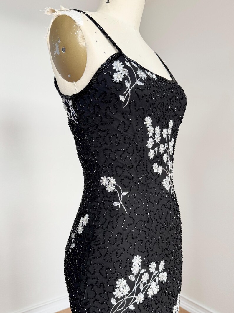 May include: Black beaded dress with white floral embroidery. The dress features spaghetti straps and a fitted silhouette. The white floral design is concentrated on the bodice and cascades down the dress. The dress is displayed on a mannequin.