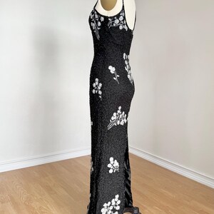 May include: A black evening gown with white floral embroidery. The dress features a fitted silhouette, spaghetti straps, and a high slit. The gown is embellished with sequins and beads, creating a shimmering effect. The dress is displayed on a mannequin.