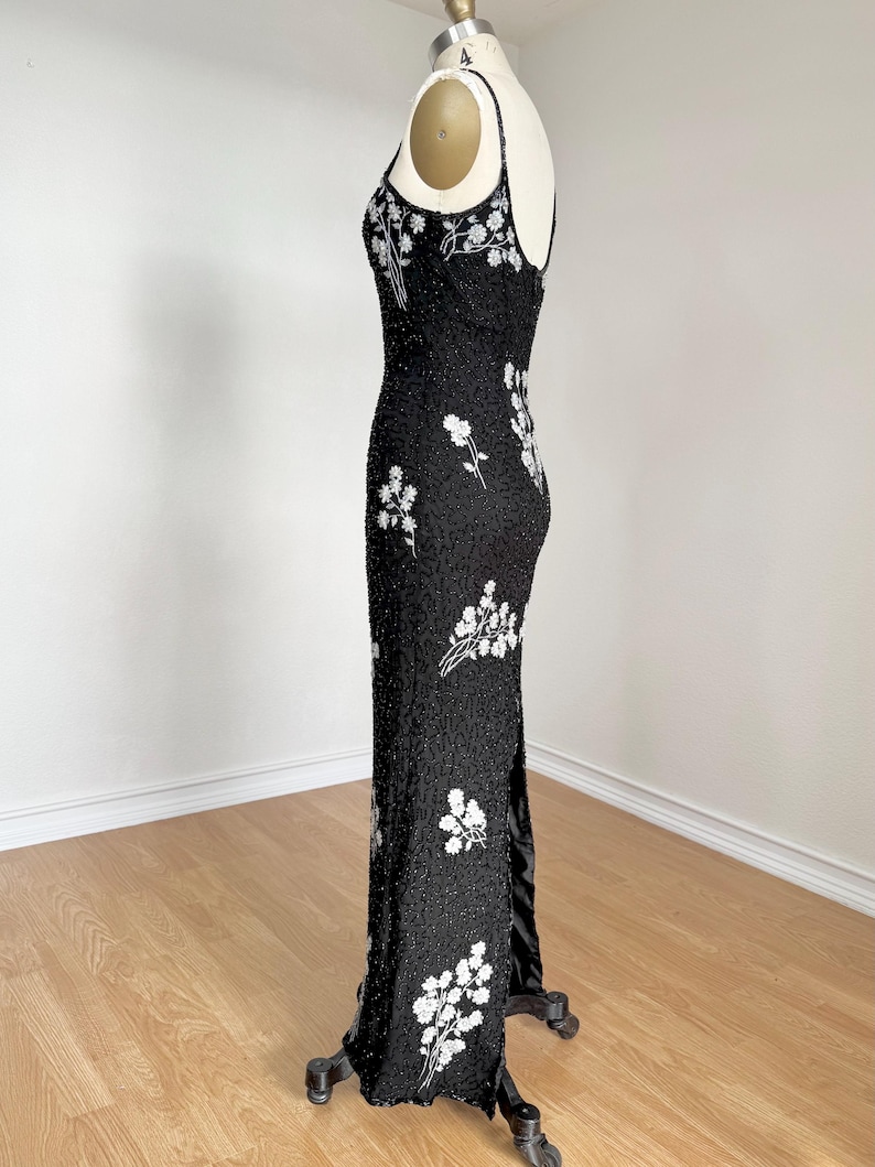 May include: A black evening gown with white floral embroidery. The dress features a fitted silhouette, spaghetti straps, and a high slit. The gown is embellished with sequins and beads, creating a shimmering effect. The dress is displayed on a mannequin.