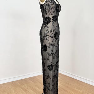 May include: A black, floor-length evening gown on a mannequin. The dress features a sheer overlay with intricate floral and leaf embroidery. The sleeveless gown has a V-neckline and a fitted silhouette, showcasing a classic, elegant design.