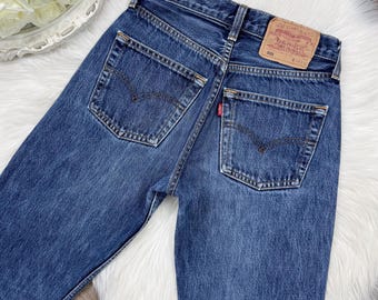 May include: Close-up of the back of blue denim jeans. The jeans have two back pockets with a curved stitch design. A small red tag is visible on one pocket. A brown leather patch with text is on the waistband.