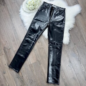 May include: Black, high-waisted, faux leather trousers with a straight leg design. The trousers have a shiny, smooth texture and a classic five-pocket style. The trousers are displayed on a wooden surface.