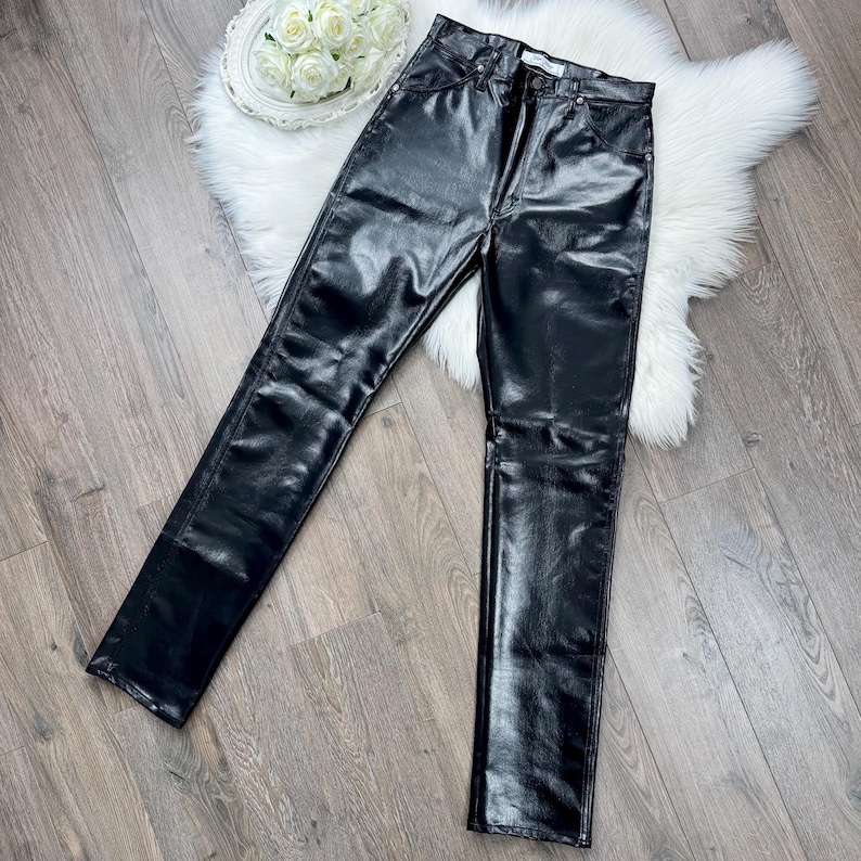 May include: Black, high-waisted, faux leather trousers with a straight leg design. The trousers have a shiny, smooth texture and a classic five-pocket style. The trousers are displayed on a wooden surface.