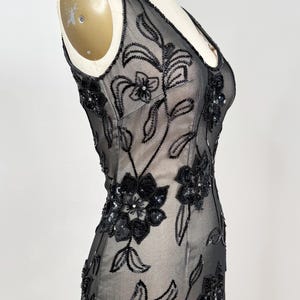 May include: A black, sleeveless evening gown with a V-neck, displayed on a mannequin. The sheer fabric is embellished with intricate floral embroidery and sequins. The dress has a fitted silhouette.