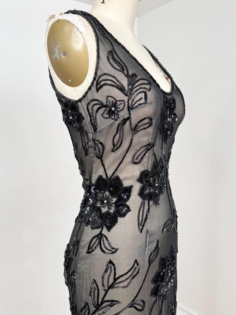 May include: A black, sleeveless evening gown with a V-neck, displayed on a mannequin. The sheer fabric is embellished with intricate floral embroidery and sequins. The dress has a fitted silhouette.