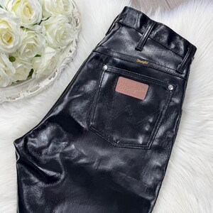 May include: Black, leather-look trousers with a classic five-pocket design. The trousers feature a Wrangler brand patch on the back pocket and a Wrangler label above the back pocket. The trousers are shiny and have a slight sheen.