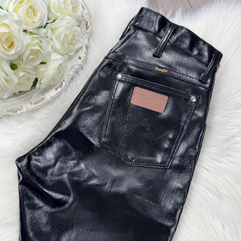 May include: Black, leather-look trousers with a classic five-pocket design. The trousers feature a Wrangler brand patch on the back pocket and a Wrangler label above the back pocket. The trousers are shiny and have a slight sheen.