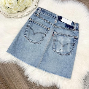 May include: Light blue denim skirt with a raw hem. The skirt features two back pockets with the Levi's logo and a small red tag. The denim has a faded look with some distressing. The skirt is displayed on a white fluffy surface.