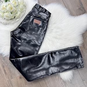 May include: Black leather trousers with a classic five-pocket design. The trousers feature a brown leather patch on the back pocket and the Wrangler logo. The trousers are laid out on a white fluffy rug.