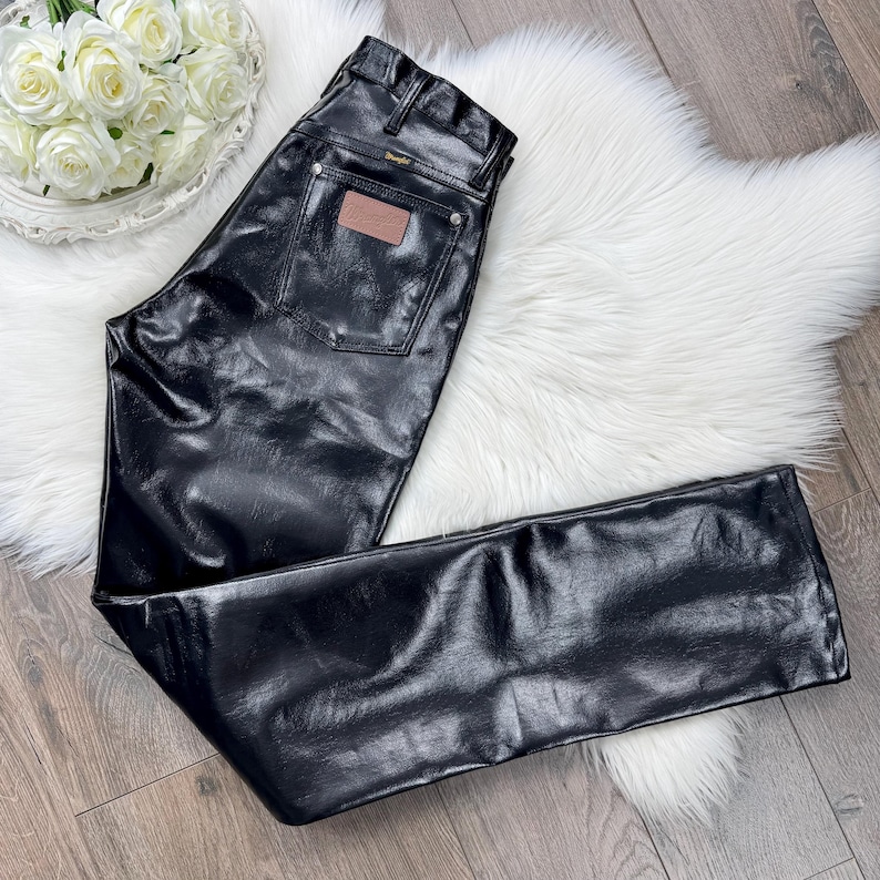 May include: Black leather trousers with a classic five-pocket design. The trousers feature a brown leather patch on the back pocket and the Wrangler logo. The trousers are laid out on a white fluffy rug.