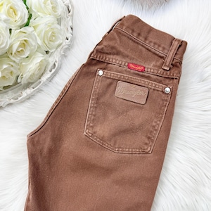May include: Brown Wrangler jeans with a high waist and a classic five-pocket design. The back pocket features a leather patch with the Wrangler logo. The jeans have a red tag and silver rivets. A stylish and timeless addition to any wardrobe.