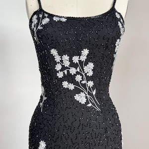 May include: A black beaded dress with white floral embroidery. The dress features thin spaghetti straps and a fitted silhouette. The floral design is concentrated on the bodice and lower portion of the dress.